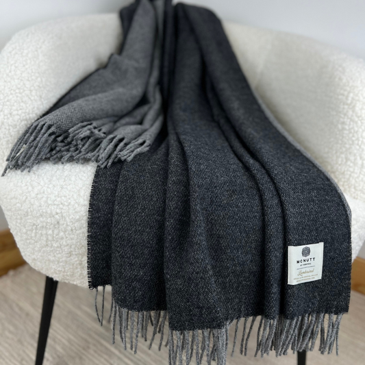 McNutt Charcoal Reversible Throw draped over a grey armchair with cushion, showing dark grey and charcoal tones